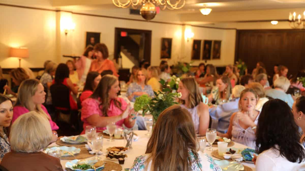 A large group of women sit at round tables set for a meal in a warmly lit dining room, engaged in conversation.