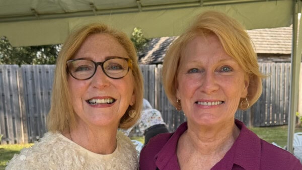 Two smiling older women with short blonde hair stand side by side outdoors under a canopy, wooden fence in the background.