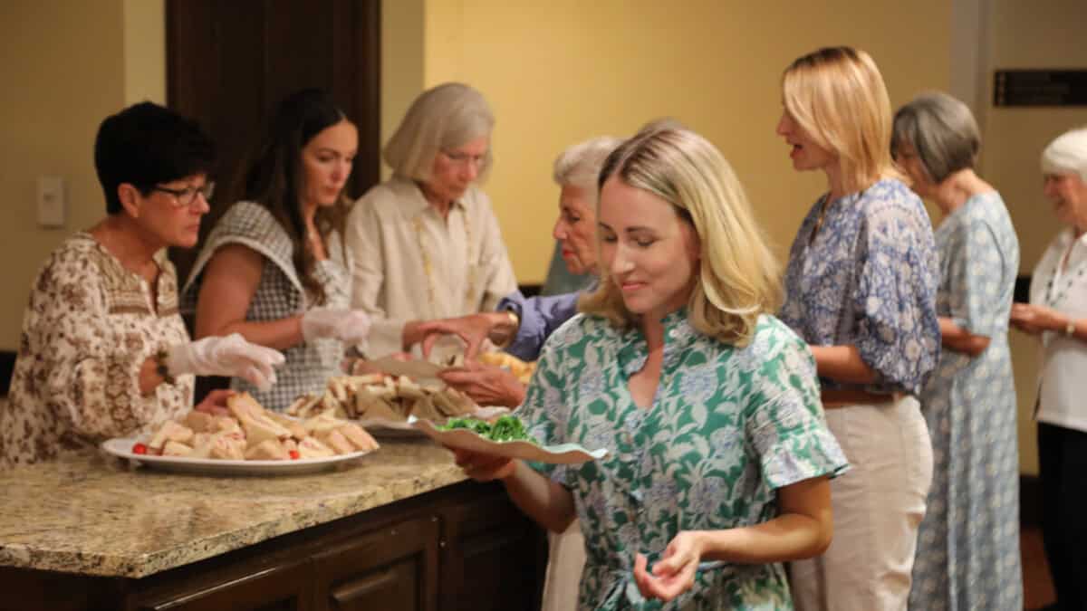 A group of women serve sandwiches and salad at a counter while a woman in front holds a plate of food.