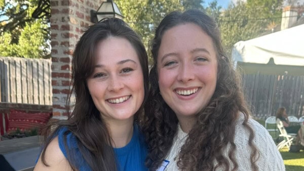 Two women smiling and standing close together outdoors, with a wooden fence and greenery in the background.
