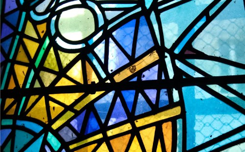 Close-up of a stained glass window with geometric patterns in blue, yellow, and green hues, intersected by black lead lines.