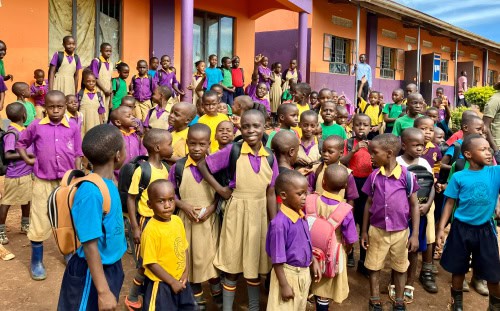 A large group of schoolchildren in uniforms stand outside a colorful school building, ready for class with Mission Partners.