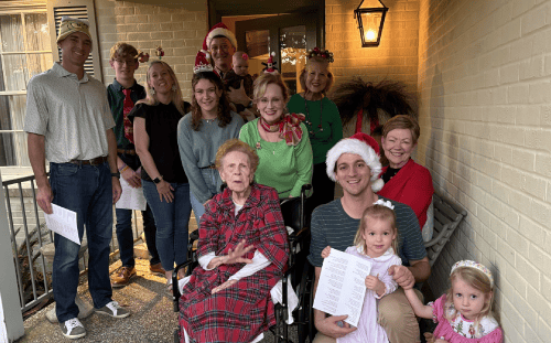 A group of people, from young children to seniors, pose on a porch, spreading Christmas joy in festive holiday attire and holding papers.