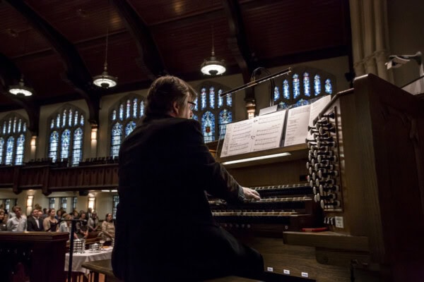 A person plays a large pipe organ in a church’s Block Library, with stained glass windows and an audience visible in the background.