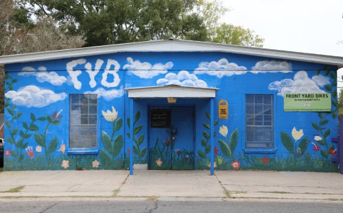 A blue building with "FYB" above, flower murals, and a "Front Yard Bikes" sign highlights the role of Mission Partners in the community.