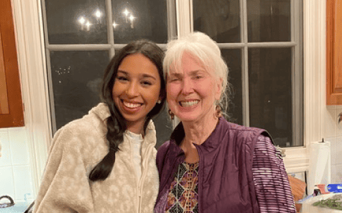 Two women stand close together in a kitchen, smiling at the camera, celebrating international friendship with cabinets and a window behind them.
