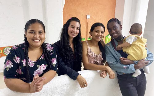 Four women stand side by side indoors, one holding a baby, all smiling as Missions Partners in front of an orange and white wall.