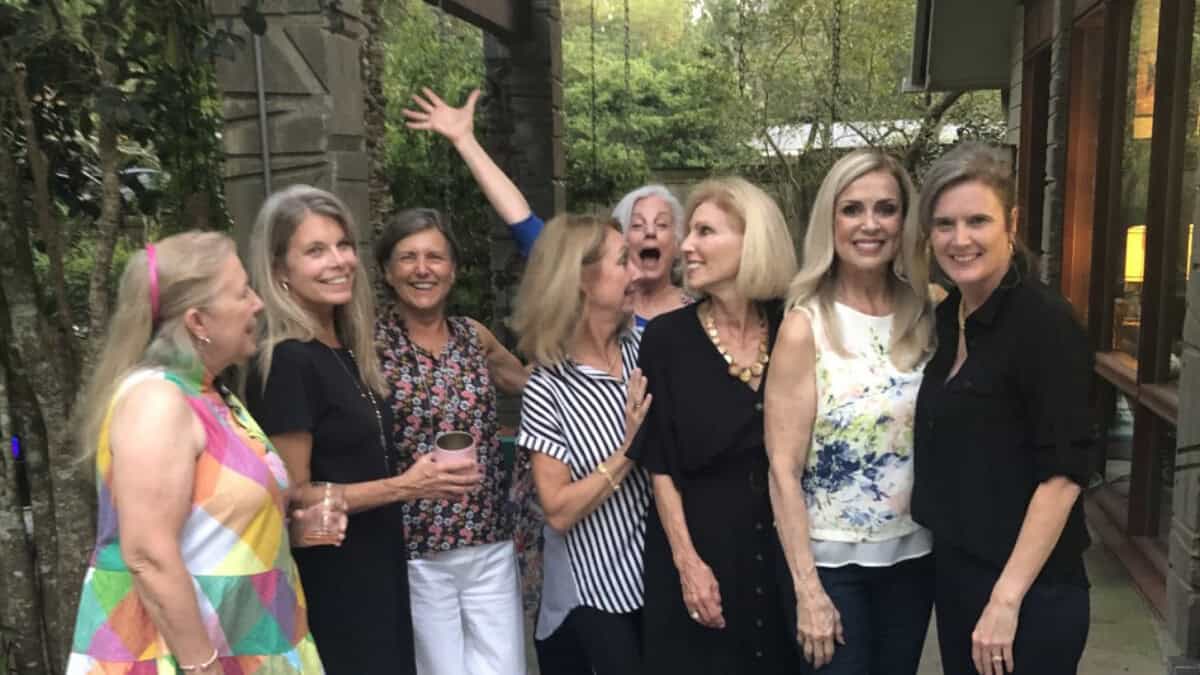 Eight women stand together outside a house, smiling and posing for a group photo in casual clothing. Trees and a building are in the background.