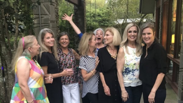 Eight women stand together outside a house, smiling and posing for a group photo in casual clothing. Trees and a building are in the background.