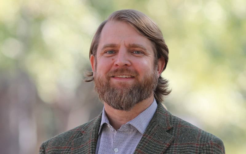 A man with a beard and mustache wearing a plaid blazer and collared shirt, standing outdoors with greenery in the background.