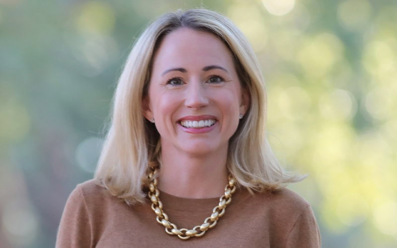 A woman with shoulder-length blonde hair, wearing a brown top and a chunky gold necklace, smiles in front of a blurred outdoor background.