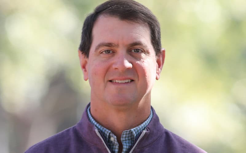 A man with short dark hair, wearing a purple jacket over a checked shirt, stands outdoors with a blurred green background.