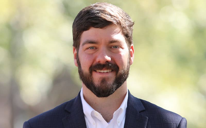 A man with short brown hair, a beard, and a dark suit jacket smiles outdoors with a blurred green background.