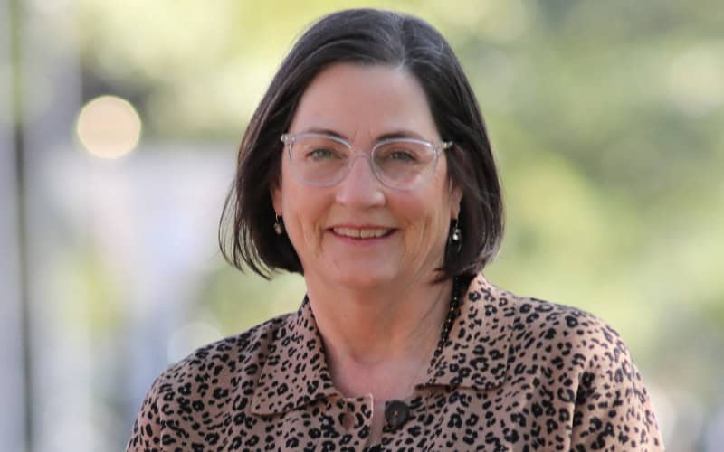 A woman with short dark hair and glasses smiles, wearing a leopard print blouse, standing outdoors with greenery in the background.
