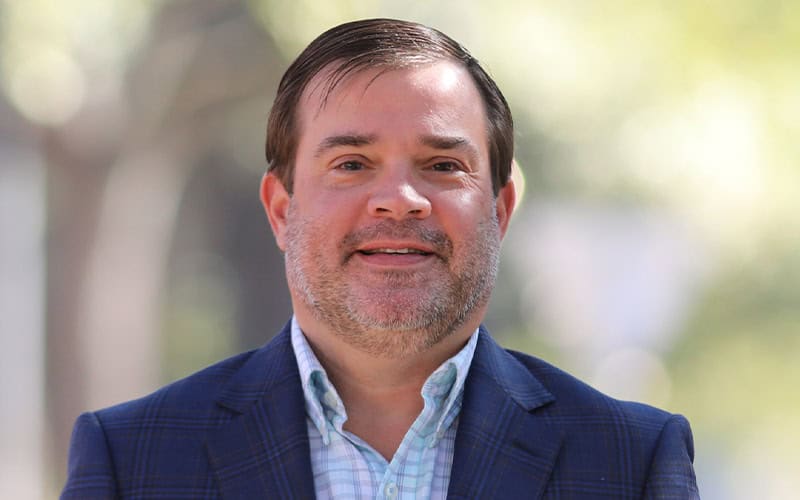 A man with short dark hair and a beard, wearing a blue checked blazer and light plaid shirt, stands outdoors with a blurred background.