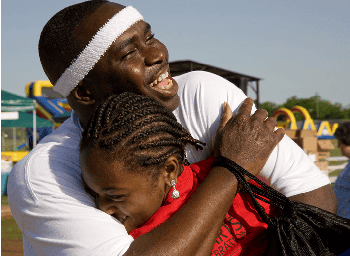 A man in a white headband hugs a smiling girl with braids at the Annual Returning Hearts Celebration, with inflatable structures behind them.