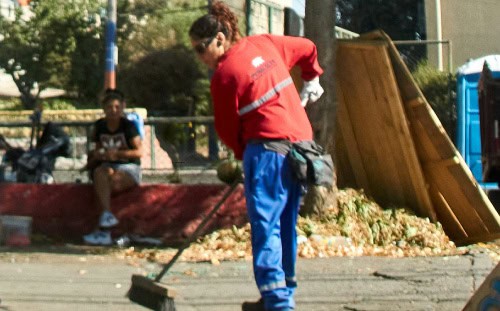 A Disaster Relief Volunteer in a red shirt and blue pants sweeps debris as two people sit on a nearby curb in the background.